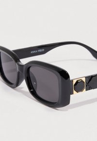 Μαύρα γυαλιά ηλίου με έντονο, τετράγωνο πλαίσιο. Τα μαύρα, υφή δακτυλίδια στα πλαϊνά έχουν χρυσή γεωμετρική λεπτομέρεια. Σκούροι φακοί με απόχρωση.