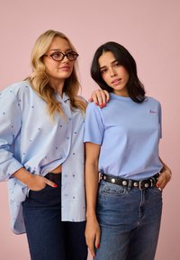 Dve ženski stojita pred rožnato ozadje; ena nosi modro srajco s srcem in očala, druga modro majico z napisom "ljubezen" in jeans.