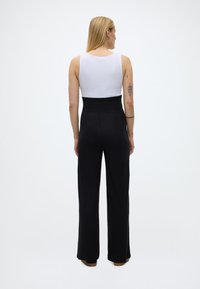 Pitkät vaaleat hiukset omaava nainen, joka pukeutuu valkoiseen hihattomaan yläosaan ja korkeavyötäröisiin mustiin leveälahkeisiin housuihin, seisoo selin yksivärisellä taustalla.