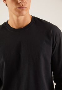 Črna majica z dolgimi rokavi iz mehkega bombažnega materiala, z okroglim izrezom, gladko teksturo in minimalističnim dizajnom.