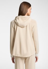 Giacca con cappuccio beige caratterizzata da una texture liscia, polsini elasticizzati e un orlo con cordino, evidenziando un design minimalista senza motivi.