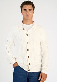 Man draagt een witte gebreide cardigan met donkere knopen over een wit overhemd, gecombineerd met een blauwe spijkerbroek, staand met één hand in de zak.