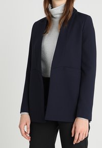 Marinblå kavaj med en strukturerad design, skräddarsydd passform och två framfickor, bärs över en silvrig polokrage. Slät textur, minimalistisk stil.