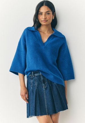 Modrý pletený svetr s výstřihem do V a oversized límcem, kombinovaný s denimovou plisovanou sukní. Textura je jemná a střih je volný.