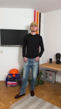 Svart långärmad tröja, blå jeans och mörka skor. Miljön inkluderar en TV, en orange lampa och ett träbord med dekorativa föremål.