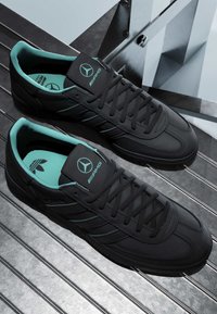 Baskets basses noires avec intérieur turquoise, logo Mercedes-Benz et AMG sur la languette, et logo Adidas sur les semelles intérieures, placées sur des escaliers en métal.