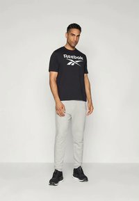 Svart kortermet Reebok t-skjorte med hvit logo, ledsaget av lysegrå joggebukser. Brukt med svarte joggesko. Enkelt bakgrunn.