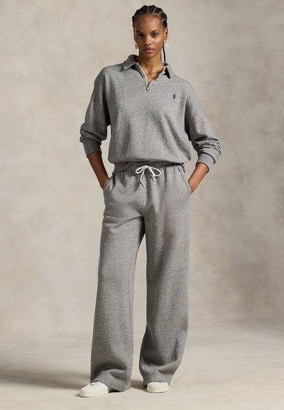 Γυναίκα που στέκεται με τα χέρια στις τσέπες, φορά γκρι ζακέτα με φερμουάρ, φαρδιά παντελόνια και λευκά παπούτσια σε ουδέτερο φόντο.