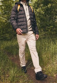 Mustat puhallinliivit, vaaleanharmaa fleece takin alla, khaki cargo-housut, mustat vaelluskengät ja keltainen reppu ruohoisessa ulkoilmassa.