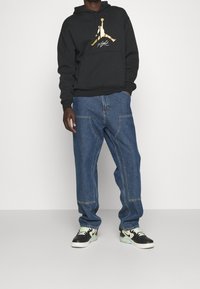 Persona che indossa un hoodie nero con il logo Jumpman dorato, jeans blu larghi con tasche frontali e scarpe nere e bianche con lacci verde chiaro.