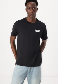 Melnas kokvilnas T-krekls ar apaļu kakla izgriezumu un balto Levi's logotipa uzlīmi uz kreisā krūša, saskaņots ar gaiši ziliem džinsiem.