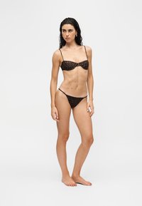 Femme aux cheveux foncés mouillés, debout pieds nus en bikini imprimé léopard sur fond blanc uni, regardant sur le côté.