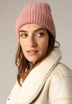 Un bonnet en tricot côtelé rose avec un motif en cable, porté avec une veste en shearling crème à col, mettant en avant la texture et la chaleur.