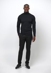 Schwarzer Rollkragenpullover kombiniert mit schwarzen Hosen und schwarzen Sneakers. Das Modell steht aufrecht vor einem schlichten weißen Hintergrund.