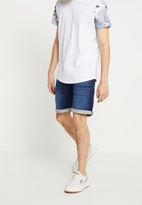 Miesten asuste, jossa on valkoinen t-paita kuvioiduilla hihoilla, tummat farkkushortsit käännetyillä reunoilla ja valkoiset lenkkarit.