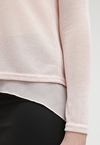 Haut à manches longues en tricot rose pâle superposé sur un chemisier transparent à ourlet asymétrique, associé à un pantalon noir ajusté sur le torse et le bras d'une personne.