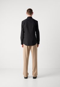 Maglietta nera a maniche lunghe con vestibilità dritta, polsini a bottone e orlo arrotondato, abbinata a pantaloni a gamba larga beige su uno sfondo neutro.