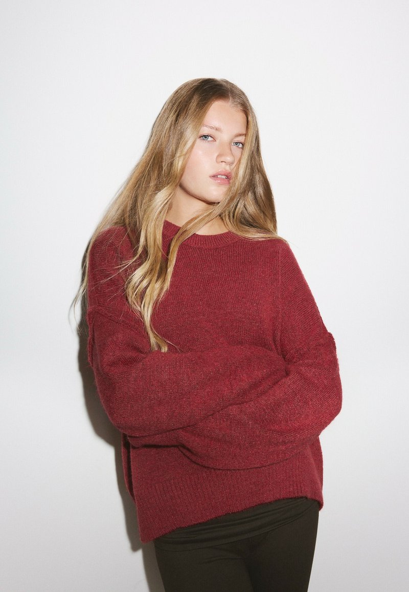 Jeune femme aux longs cheveux blonds portant un pull rouge et un pantalon noir, debout les bras croisés devant un fond blanc.