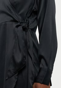 Vestido envolvente em satin preto com mangas longas, detalhe de cintura amarrada, textura suave e decote sem gola.