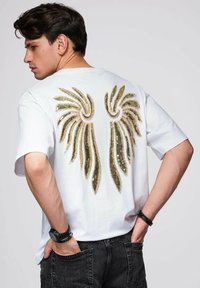 Biała koszulka z krótkim rękawem o luźnym kroju, wykonana z jednolitego koloru materiału, z dużym złotym wzorem skrzydła z rhinestones na plecach.