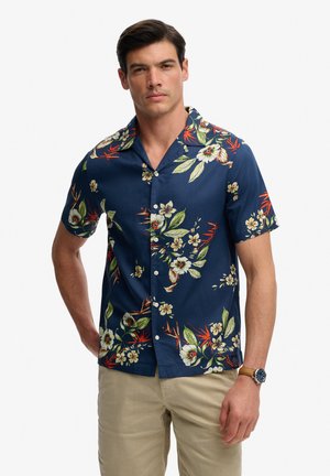 Uomo che indossa una camicia blu navy a maniche corte con bottoni, con stampe floreali bianche e arancioni, pantaloni beige e un orologio da polso.