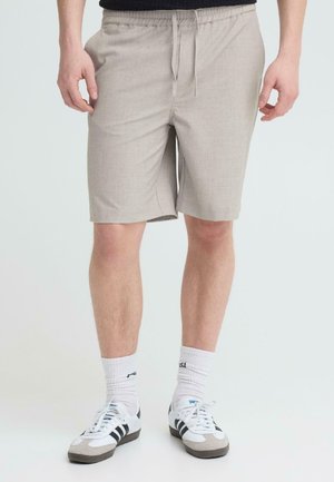 Mees, kes kannab beežikat paelaga šorte, valgeid põlvesokke ja valgeid tossusid mustade triipudega, üksluisel taustal.