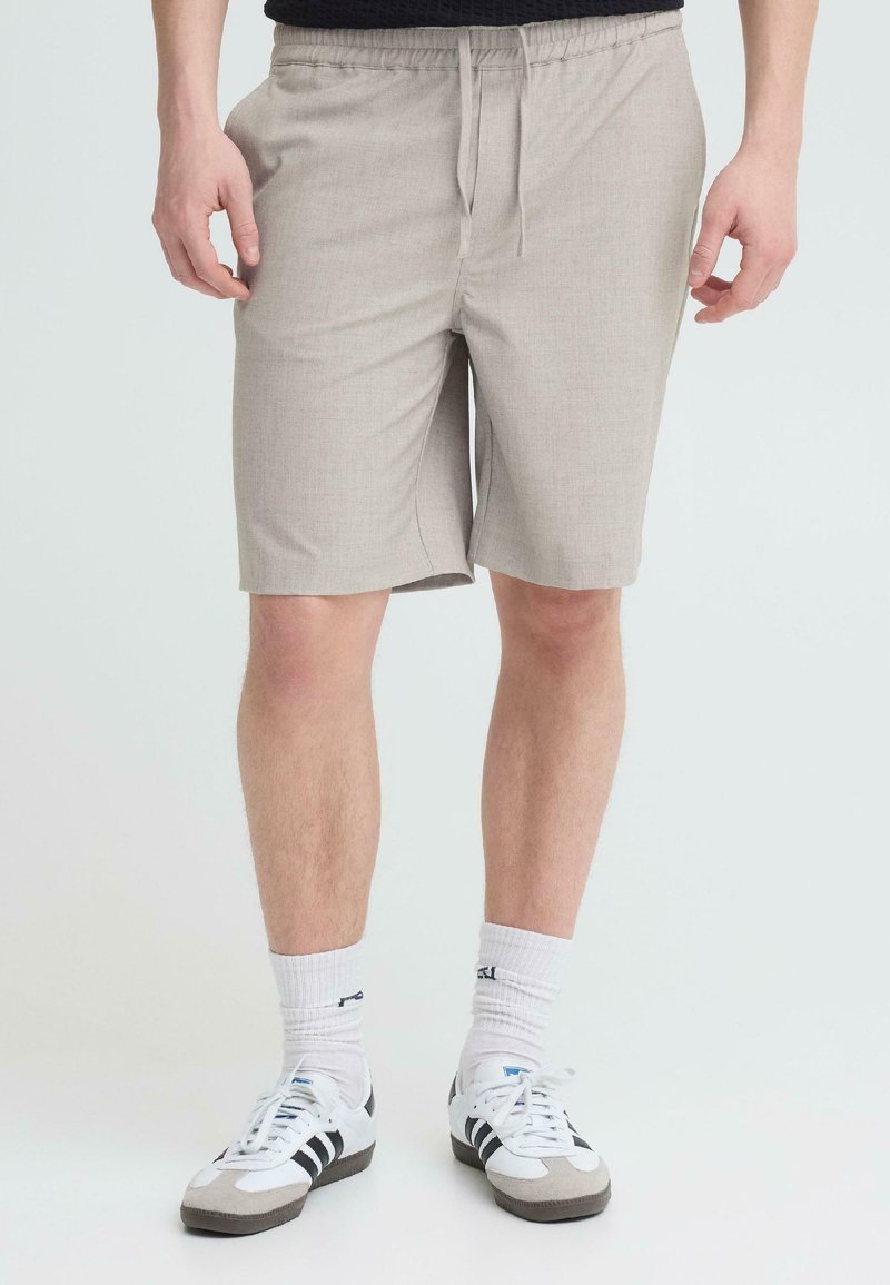 Άνδρας που φοράει μπεζ σορτς με κορδόνι, λευκές κάλτσες μέχρι τον αστράγαλο και λευκά αθλητικά παπούτσια με μαύρες ρίγες σε απλό φόντο.