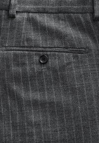 Gros plan sur un tissu gris à fines rayures avec une petite poche passepoilée boutonnée et des passants de ceinture visibles sur un pantalon sur mesure.