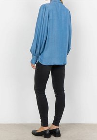 Camicia leggera blu chiaro a maniche lunghe con spalle a palloncino, abbinata a jeans skinny neri e scarpe basse nere. Tessuto morbido, design casual.