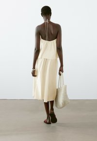 Robe sans manches jaune clair avec un lien à l'arrière, design à couches et coupe ample. Porte un sac à main en osier et des sandales noires.