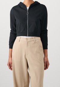 Mujer vistiendo una sudadera corta con cremallera negra y bolsillos frontales, junto con pantalones beiges de tiro alto con botón y pasadores para cinturón.