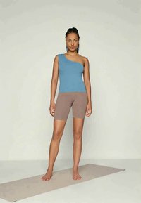 Sieviete stāv basām kājām uz jogas paklāja, ģērbusies zilā pleca topā un bēšās velo šortos, fona krāsa ir neitrāla.