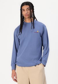 Chemise en coton bleu à manches longues avec un col rond. Présente un petit logo de marque orange et rouge sur la poitrine et une coupe décontractée.
