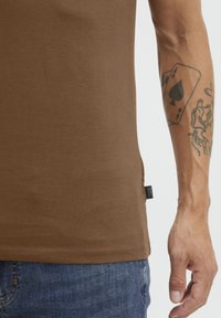 Ruda trumpa rankovė t-shirt, pagaminta iš minkštos medžiagos, su šoniniu etiketu ir priartinta nuotrauka, kurioje matoma ranka su matomu tatuiruotė ir džinsai.