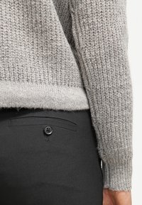 Gros plan d'une personne portant un pull en tricot texturé gris et un pantalon noir avec une poche arrière boutonnée.