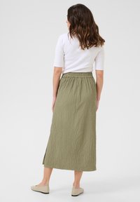 Olijfgroene, gestructureerde lange rok met elastische tailleband. Gecombineerd met beige ballerina's, gezien vanaf de achterkant.