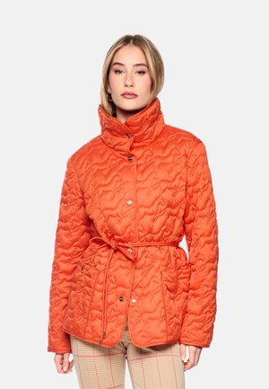 Frau trägt eine leuchtend orange gesteppte Jacke mit hohem Kragen und gebundener Taille, kombiniert mit hell karierten Hosen.