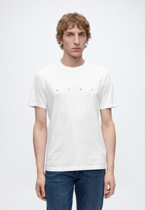 Ung mand med lyst brunt hår iført en ensfarvet hvid T-shirt med små blå "HUGO" bogstaver og blå jeans, stående mod en hvid baggrund.