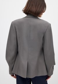 Pilkas sportinis švarkas su struktūrizuota dizainu, su vienu ventiliaciniu angos gale, plačiomis atvartais ir tekstūruotu audiniu. Šiek tiek per didelis modelis.