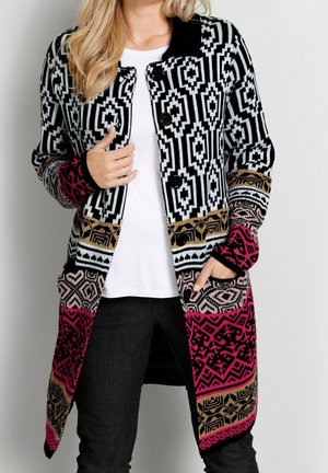 Cardigan long en tricot avec des motifs géométriques noirs, blancs, roses et beiges, boutonné à l'avant, porté sur un haut blanc et un pantalon noir.