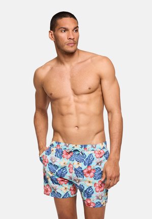 Hombre sin camiseta que lleva pantalones cortos de baño florales y coloridos con las manos en los bolsillos, mirando hacia un lado contra un fondo liso.