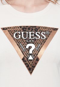 Logo GUESS con stampa leopardata e borchie in bronzo su una maglietta bianca, con le parole "WASHED JEANS" e "U.S.A." all'interno di un triangolo.