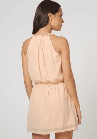 Vestido sin mangas de color melocotón con escote fruncido, cintura elástica y tela suave y fluida; cae por encima de la rodilla con un sutil dobladillo semitransparente.