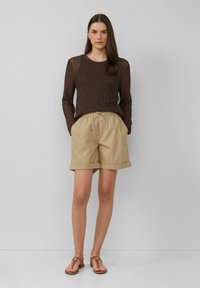 Pull en maille côtelée marron, short beige avec les manches repliées, et sandales marron. Tissu léger, coupe décontractée. Le modèle se tient debout les mains dans les poches.
