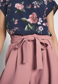 Floral patterned navy top with short sleeves above light pink high-waisted trousers featuring a bow tie at the waist and pleated design.