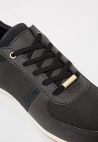 Svart sneaker med strukturerat tyg, platta snören, guldaccenter och vit sula. Har släta och ribbade material för kontrast.