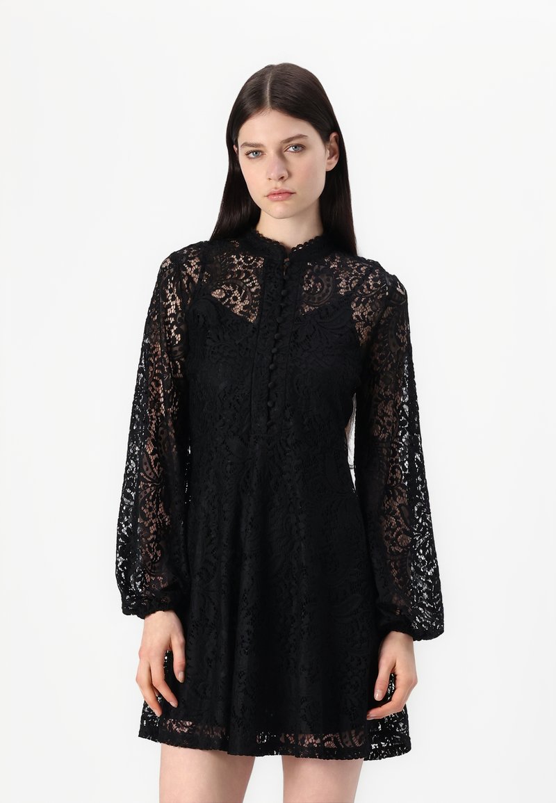 Schwarzes Spitzenkleid mit langen, gepufften Ärmeln, einem hohen Kragen und Knopfdetails an der Vorderseite. Besitzt ein filigranes Blumenmuster.