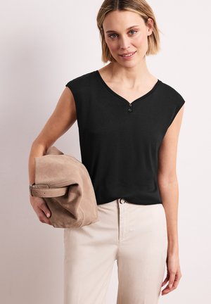 Mujer de cabello corto que lleva una blusa negra sin mangas y pantalones claros, sosteniendo una chaqueta de ante beige doblada en una mano.