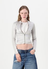 Světlá šedá zkrácená bunda se zapínáním na zip, hladkým povrchem a dlouhými rukávy, kombinovaná s džíny z modrého denimového materiálu s vysokým pasem.