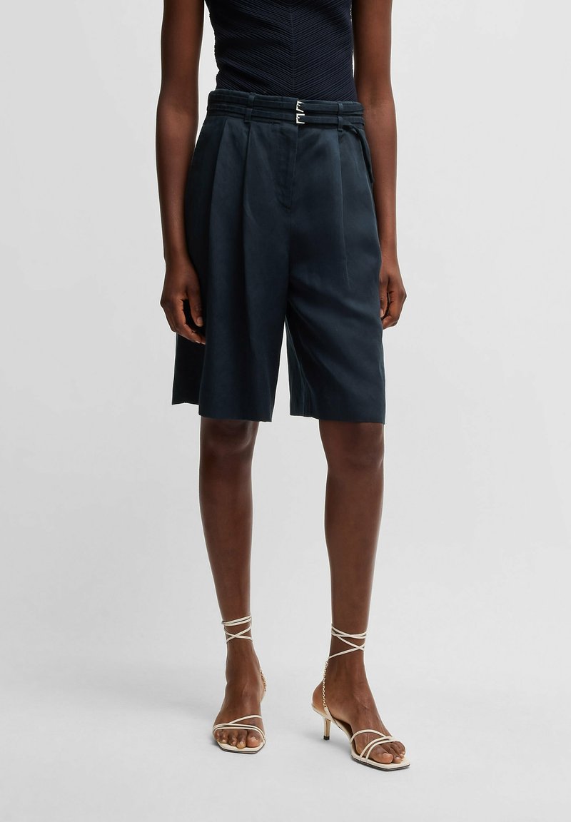 Femme portant un short ample bleu marine taille haute avec ceinture à double boucle et des sandales blanches à talons et à brides sur fond uni.